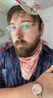 A selfy photo of me, Seth Thill, in a Barbie-themed bedroom. I am wearing transparent-framed glasses, a navy and red floral shirt, a pink floral kerchief, and a white and pink floral baseball cap. 