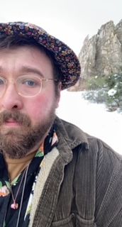 A selfy photo of me, Seth Thill, standing in front of a snow-covered rock formation at the Garden of the Gods in Colorado Springs, Colorado. I am wearing transparent glasses, a floral shirt, brown jacket, and floral bucket hat.