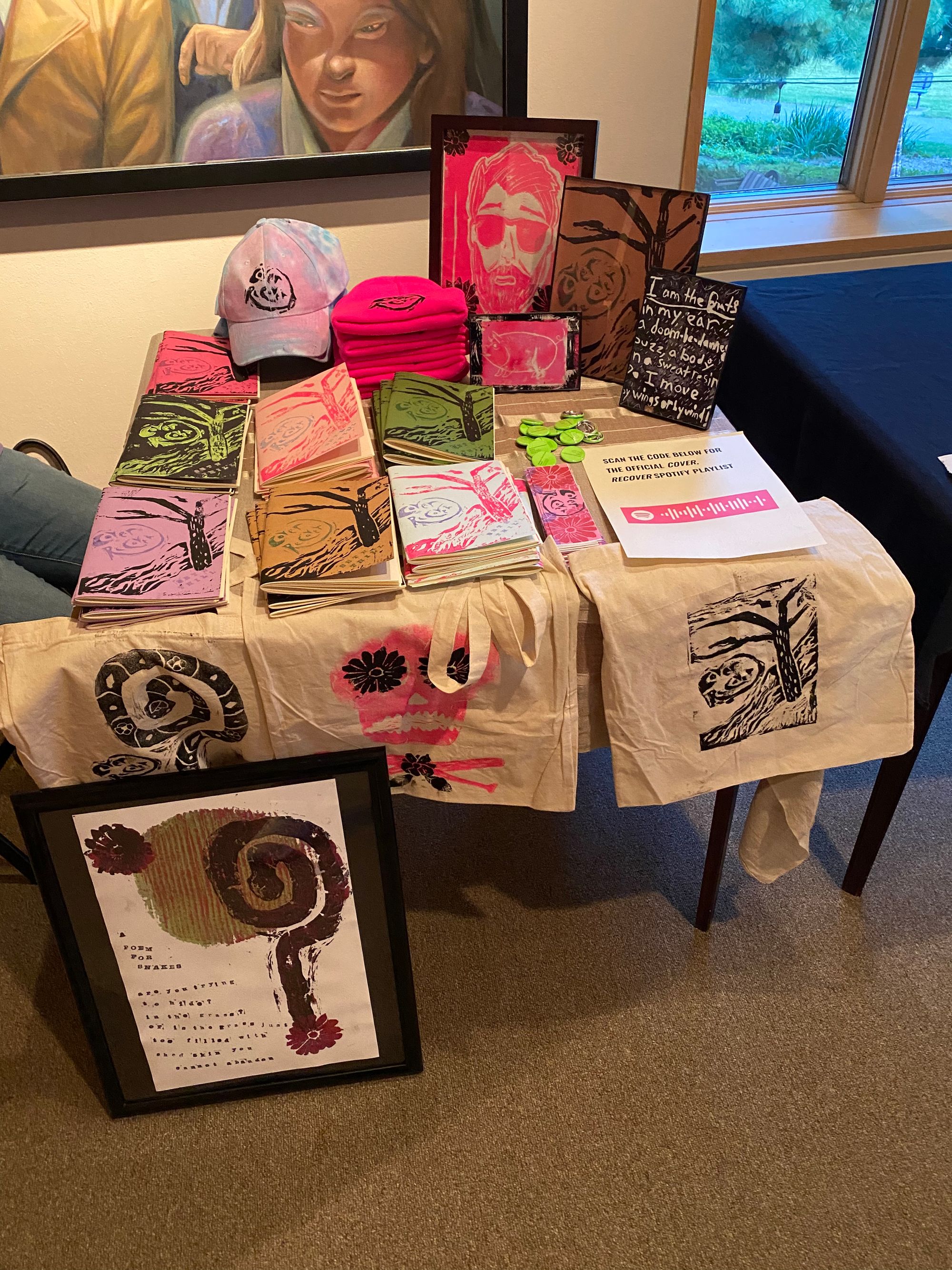 A table filled with copies of the chapbook, Cover, Recover, by Seth Thill, and merch with artwork related to the book.There are blue and pink tie-dyed ballcaps with the book titled printed on them, pink stocking caps with the title printed on the front, and five linocut carvings.