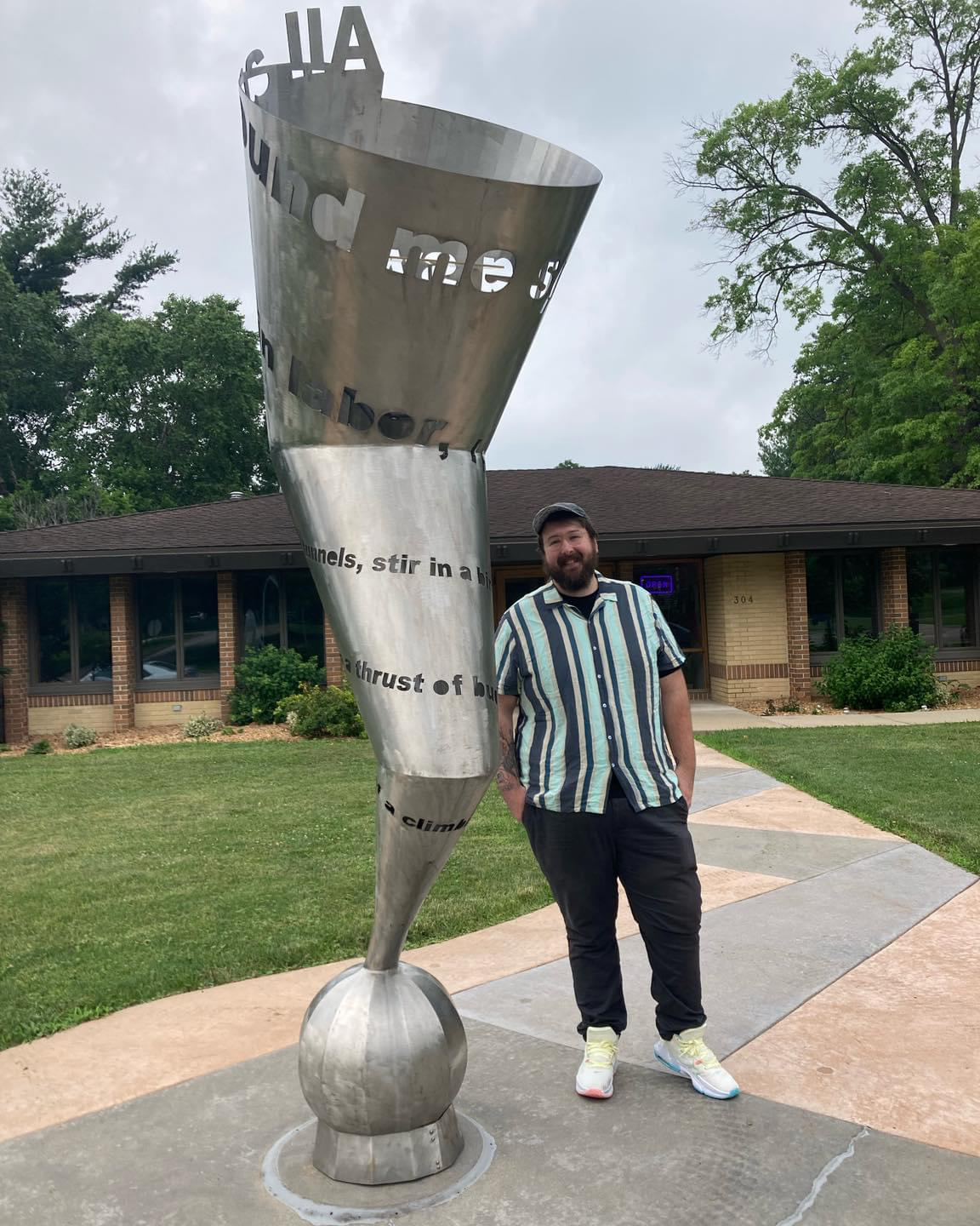 Seth Thill standing outside of the Hearst Center in Cedar Falls Iowa, next to a large (10-15 feet tall) metal statue of a horn. Seth is wearing a vertically striped camp shirt with multiple shades of blue, grey pants, a baseball cap, and white Nikes. 