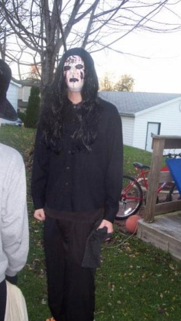 14 year-old Seth standing in his backyard in a homemade Joey Jordison costume. He is wearing black pants and a black button down shirt, a wig of long, straight, black hair, and an expressionless Kabuki mask painted with black lines, clay faux stitches, and fake blood.