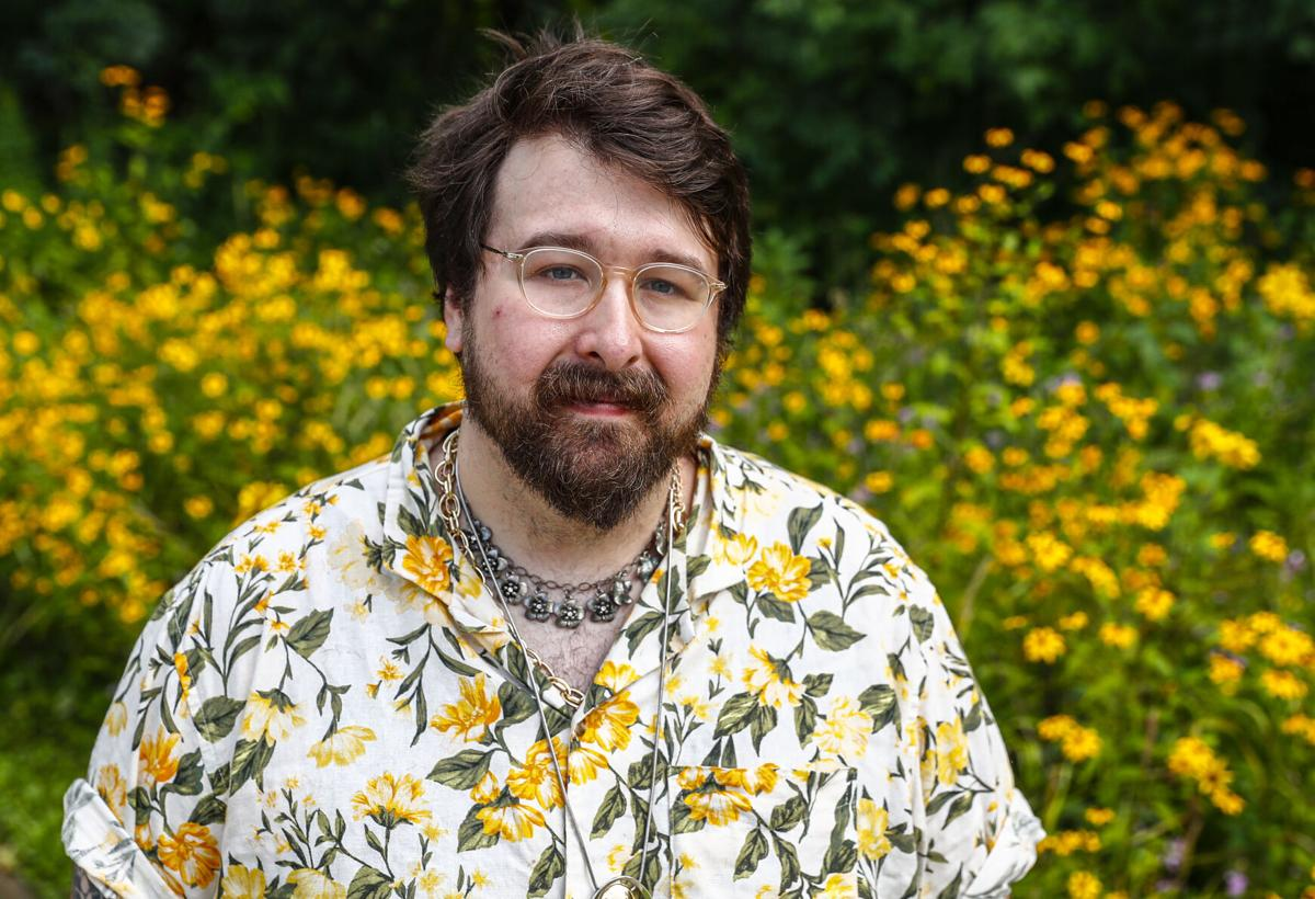 Bearded man with clear-framed glasses, silver chain necklace, and a yellow floral camp shirt stands in front of a field of yellow wildflowers. 